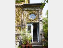 Fassade eines historischen Hauses mit Efeubewuchs, einem runden Fenster und einem Eingang mit verzierten Glastüren. Vor der Treppe stehen mehrere Topfpflanzen, und rechts davon ist eine steinerne Figur auf einem Sockel sichtbar.