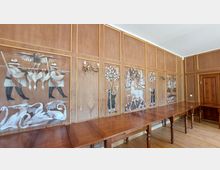 Holzvertäfelter Raum mit Wandmalereien, die Jagdszenen und Tiere wie Schwäne, einen Hirsch und Vögel darstellen. Der Raum ist mit langen Holztischen und einem Kronleuchter an der Wand ausgestattet.