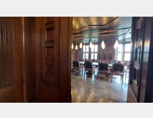 Ein historischer Tagungsraum mit holzgetäfelten Wänden, großen Fenstern mit Sprossen und einem langen Konferenztisch, der von Holzstühlen umgeben ist. Von der Decke hängen mehrere Pendelleuchten, und im Hintergrund sind Regale sowie weitere dekorative Holzelemente sichtbar.