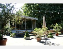Ein Biergarten mit Holzbänken und Tischen, umgeben von Pflanzgefäßen mit Sträuchern und Blumen, unter freiem Himmel im Schatten von großen Bäumen. Im Hintergrund ist ein Gebäude mit großen Glasfenstern zu sehen.