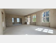 Unmöblierter, renovierungsbedürftiger Innenraum mit glatten, verputzten Wänden und hellem Boden. Mehrere große Sprossenfenster sowie eine Glastür lassen viel Tageslicht hinein und bieten Blick ins Grüne.