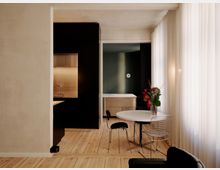 Modernes, minimalistisches Wohninterieur mit hellem Holzboden, einer schwarzen Küchenzeile mit Edelstahl-Nische und einem runden weißen Esstisch. Rechts sorgen bodentiefe, halbtransparente Vorhänge für Tageslicht; im Hintergrund ist eine Schlafnische mit Bett und dunkler Akzentwand zu sehen.