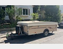 Campinganhänger in beiger Farbe mit flachem, geschlossenen Dach, abgestellt am Straßenrand vor einem Wohnhaus mit Gartenzaun und Büschen. Im Hintergrund sind Häuser mit weißen Fassaden sichtbar.