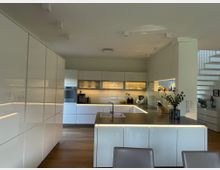 Moderne Küche mit glänzenden, weißen Einbauschränken und einer zentralen Kücheninsel mit grauer Arbeitsplatte und integriertem Waschbecken. Helle LED-Beleuchtung unter den Schränken und der Insel sorgt für indirektes Licht, während eine offene Treppe und Dekorationsgegenstände im Hintergrund sichtbar sind.