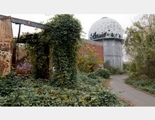 Verlassenes Gelände mit überwucherten Strukturen, darunter eine mit Graffiti bedeckte Mauer und überwachsene Metallgerüste. Im Hintergrund steht eine geodätische Kuppel, die teilweise beschädigt ist, sowie eine asphaltierte Gehwegfläche, die durch das wild bewachsene Areal führt.