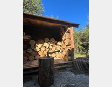 Offener Holzunterstand im Freien, gefüllt mit gestapelten Baumstämmen und Rundhölzern unter einem flachen Dach. Davor steht ein Hackklotz mit einem Beil; umgeben von Kiesboden, Büschen und Bäumen unter blauem Himmel.