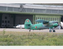 Ein grünes Doppeldeckerflugzeug steht auf einer asphaltierten Fläche vor einem großen Hangar mit grünen Toren. Im Hintergrund sind weitere Flugzeuge und Gebäude sichtbar.