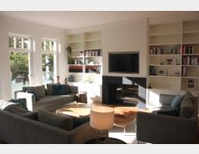 Helles Wohnzimmer mit großen Fenstern, die einen Blick auf einen Garten bieten. Der Raum enthält zwei graue Sofas, einen Holztisch mit Schachbrett, eine schwarze Kaminumrandung mit darüber aufgehängtem Fernseher sowie Bücherregale an der Wand.