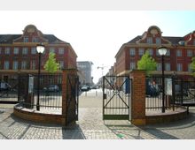 Eingangsbereich mit einem offenen, schmiedeeisernen Tor und Ziegelmauern, dahinter eine gepflasterte Straße, die zwischen symmetrischen, roten Backsteingebäuden mit verzierten Giebeln und mehreren Fenstern verläuft. Im Hintergrund sind Autos, Menschen und ein Baukran sichtbar.