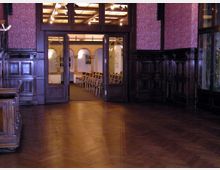 Holzvertäfelter Raum mit glänzendem Parkettboden, einer offenen Tür, die zu einem hell beleuchteten Nebenraum führt, in dem Stuhlreihen aufgestellt sind. Der Nebenraum ist durch einen Türrahmen mit Glasverzierungen und Säulen sichtbar.