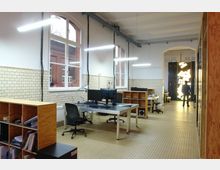 Großzügiges, modernisiertes Büro in einem ehemaligen Industriegebäude mit hohen Decken, Fliesenwänden aus gelblichem Backstein und großen Fenstern mit weißen Rahmen. Der Raum ist mit Schreibtischen, Bürostühlen und Regalen ausgestattet, während eine leuchtende, geometrische Beleuchtung im Hintergrund zu sehen ist.