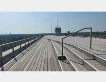 Große Holzterrasse mit Geländern aus Metall und Drahtnetz, im Hintergrund ein technisches Gebäude mit Antennen. Die Terrasse bietet einen weiten Blick auf eine städtische Umgebung mit Bäumen und vereinzelten Hochhäusern am Horizont.