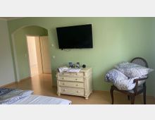 Ein Schlafzimmer mit hellgrünen Wänden, einem an der Wand befestigten Flachbildfernseher, einer beigefarbenen Kommode und einem Holzstuhl mit darauf gestapelter Bettwäsche; im Hintergrund ist eine offene Tür zu sehen, die in einen anderen Raum führt.