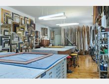 Werkstatt für Bilderrahmen mit einer Wand voller Rahmen unterschiedlicher Größen und Farben, langen Holzleisten in der Ecke, großen Arbeitstischen in der Mitte und Regalen mit Werkzeugen und Materialien an der Seite. Der Holzboden und helle Leuchtstofflampen schaffen eine funktionale Arbeitsumgebung.