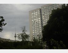 Ein Hochhaus mit zahlreichen Fenstern und Balkonen steht vor einem bewölkten Himmel. Im Hintergrund ist der Berliner Fernsehturm mit seiner markanten Kugel und Antennenspitze sichtbar, umgeben von Bäumen im Vordergrund.