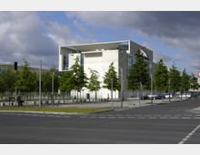 Modernes Regierungsgebäude mit kantiger Architektur, hellen Betonfassaden und großen Glasflächen, umgeben von jungen, symmetrisch gepflanzten Bäumen und breiten Gehwegen; eine Straßenkreuzung ist im Vordergrund sichtbar.