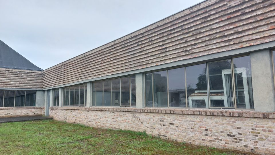 Flaches Gebäude mit moderner Architektur, bestehend aus einer Fassade aus Ziegelstein und großen Fensterfronten mit schmalen Rahmen. Im Vordergrund befindet sich eine Rasenfläche, während der Himmel wolkenverhangen ist.