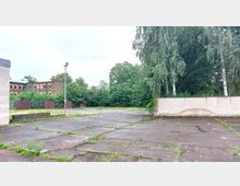 Ein leerer, verwitterter Parkplatz mit vereinzeltem Pflanzenbewuchs und einer niedrigen Backsteinmauer im Hintergrund. Dahinter befinden sich zahlreiche Bäume sowie ein älteres Backsteingebäude.