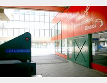 Eingangsbereich des U-Bahnhofs Schlossstraße mit einer rolltreppenartigen Struktur und einem blauen Schild. Daneben ein modernes Gebäude mit roten Fassadenplatten, großen Fenstern und Beschriftungen wie „Spielwaren“, „Basteln“ und „Bistro“.