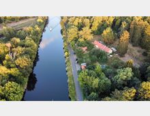 Luftaufnahme einer Waldlandschaft mit einem schmalen Fluss, der von Bäumen gesäumt ist, und einem Boot darauf. Rechts des Flusses befindet sich ein Gebäude mit roten Dächern, umgeben von einem Garten, einem kleinen Pool und parkenden Autos.