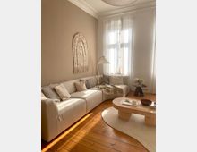 Helles Wohnzimmer mit einer cremefarbenen Eckcouch, dekoriert mit Kissen und einer Decke. Im Vordergrund steht ein beiger Couchtisch auf einem runden Teppich, dahinter ein großes Fenster mit transparenten Vorhängen, das den Raum mit Tageslicht erfüllt.