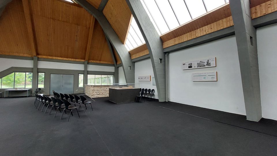 Moderner Saal mit hohen, freitragenden Holzdecken und Betonstützen. Der Raum enthält eine Sitzreihe mit schwarzen Stühlen, einen kleinen Infotresen aus Stein und Glas sowie Informationsplakate an einer weißen Wand; große Fenster lassen Tageslicht herein.