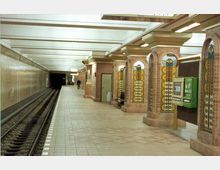 Unterirdische U-Bahn-Station mit verzierten Backsteinpfeilern, die mit bunten Glas- und Fliesenmustern dekoriert sind. Links sind die Gleise zu sehen, während rechts ein Fahrkartenautomat und Sitzbänke entlang des Bahnsteigs stehen.
