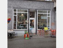 Außenansicht eines kleinen Ladens mit grauer Fassade und großen Fenstern. Vor dem Schaufenster stehen bunte Tische und Stühle, dazu dekorative Gegenstände wie ein Spiegel und eine grüne Glasvase; Schilder am Fenster informieren über Öffnungszeiten und Liefermöglichkeiten.