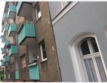 Mehrfamilienhaus mit brauner Fassade und türkisfarbenen Balkonen sowie einem neu gestrichenen grauen Teilbereich mit weiß gerahmtem Rundbogenfenster im Erdgeschoss. Einige Balkone sind mit Blumenkästen dekoriert.