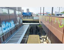 Blick auf Gleise einer Bahnstation, eingerahmt von zwei braunen Gebäudewänden mit Graffiti und einer Brücke im Hintergrund, über der Fahrzeuge und Personen sichtbar sind. Im Hintergrund sind moderne Hochhäuser und der Berliner Fernsehturm zu erkennen.