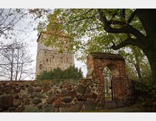 Alter Kirchturm aus Naturstein und Fachwerk, umgeben von einer Mauer aus unregelmäßigem Feldstein. Im Vordergrund ein gemauerter Torbogen mit schmiedeeisernem Gitter, umgeben von Bäumen mit Herbstlaub.
