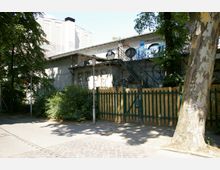 Ein verwittertes Gebäude mit Graffiti an den Wänden, einer Metalltreppe an der Seite und einem grünen Holzzaun im Vordergrund. Die Umgebung ist von Gehwegen, Bäumen und Büschen geprägt.