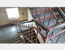 Eine abgenutzte Innentreppe in einem Gebäude mit verwittertem Metallgeländer und abblätternder Farbe. Im Hintergrund sind ein großes Fenster, eine Heizung und eine Wand mit sichtbaren Gebrauchsspuren zu sehen.