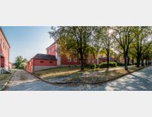 Straßenszene mit roten Wohngebäuden in einem ruhigen Viertel, umgeben von Bäumen. Ein gepflasterter Weg verläuft entlang der Häuser, und ein kleiner Grünstreifen trennt Gehweg und Gebäude.