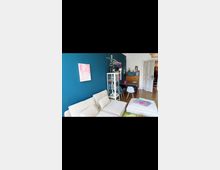 Wohnzimmer mit einer cremefarbenen Sofaecke und einer blauen Wand, an der ein abstraktes Gemälde hängt. Im Hintergrund stehen ein weißes Regal mit Gläsern, ein Holztisch mit Stühlen und eine halboffene Tür, die in einen weiteren Raum führt.