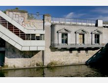 Außenansicht eines Gebäudes an einem Gewässer mit einer gemauerten Fassade und mehreren klassischen Fenstern mit Dreiecksgiebeln. Links unten verläuft eine schräg angebrachte Metalltreppe mit Glasfenstern, und an der Wand ist Graffiti zu sehen.