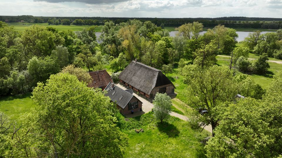 Luftaufnahme eines ländlichen Hofes mit mehreren Gebäuden, darunter eine große Scheune mit Reetdach und kleinere Häuser mit Ziegeldach, umgeben von Wiesen und dichtem Baumbestand. Im Hintergrund sind ein Flusslauf sowie weite Felder unter einem wolkigen Himmel zu sehen.