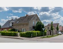 Einfamilienhaus in einer Wohnsiedlung, gekennzeichnet durch eine Fassade in Braun- und Beigetönen, ein Satteldach und weiße Fensterrahmen; vor dem Haus befindet sich eine Hecke mit einem kleinen Gartentor. Im Hintergrund sind weitere Häuser mit roten Ziegeldächern und ein Gehweg entlang der Straße zu sehen.