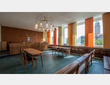 Innenraum eines Besprechungsraums mit Holzmöbeln, darunter Tische und Bänke, sowie einer mit Holz verkleideten Rückwand. Große Fenster mit bunten vertikalen Vorhängen lassen Tageslicht herein, im Hintergrund ist der Turm eines Gebäudes zu sehen.