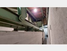 Industriehalle mit einer großen grünen Metallkonstruktion, die sich entlang einer Betonschiene erstreckt. Über der Konstruktion ist ein Holz- und Metallgebälk sichtbar, während die Wände grau verputzt sind.