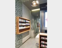Innenansicht einer modernen Apotheke mit minimalistischer Gestaltung. An der Wand hängen Regale aus Holz mit Pflege- und Gesundheitsprodukten, die vor einer Mosaikwand angebracht sind; daneben stehen schlichte Waschbecken aus Beton.