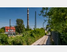Schmaler Weg mit grünem Bewuchs auf beiden Seiten, links ein zweistöckiges Haus mit rotem Dach, daneben Strommasten und ein Schornstein in der Ferne. Im Hintergrund sind Bäume und ein blauer Himmel zu sehen.