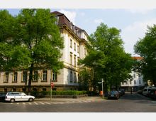 Historisches Wohn- oder Verwaltungsgebäude mit heller Fassade und großen Fenstern, umgeben von grünen Bäumen. Im Vordergrund eine gepflasterte Straßenecke mit parkenden Autos und einem Gehweg.