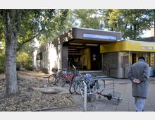 Eingang zum U-Bahnhof Halemweg mit überdachtem Eingangsbereich, einem danebenliegenden gelben Imbissstand und mehreren abgestellten Fahrrädern im Vordergrund. Im Hintergrund sind Bäume und ein schattiger Gehweg sichtbar.