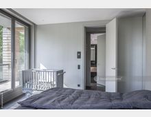 Modernes Schlafzimmer mit großen Fenstern, die auf einen Balkon blicken. Im Raum stehen ein graues Bett und ein Kinderbett, während eine geöffnete Tür den Blick in ein angrenzendes Badezimmer freigibt.