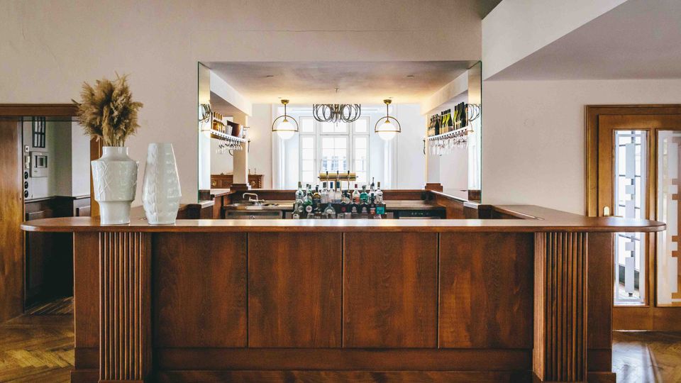 Barbereich mit einer Theke aus dunkelbraunem Holz, dekoriert mit weißen Vasen und getrockneten Pampasgräsern. Im Hintergrund sind Hängeregale mit Gläsern, eine Spiegelwand sowie große Fenster und moderne Hängelampen zu sehen.