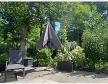 Garten mit einer gepflasterten Terrasse, auf der ein grauer Sonnenschirm und ein Gartenstuhl stehen. Umgeben von dichtem Grün, blühenden Pflanzen in Kübeln und hohen Bäumen im Hintergrund.