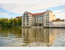 Großes mehrstöckiges Ufergebäude mit weißer Fassade, dunklen Fachwerk-Elementen und roten Ziegeldächern direkt am Fluss, mit vielen Fenstern und einem kleinen Anleger am Wasser. Links grenzt eine Baumreihe ans Ufer, rechts ist ein Gebäudeteil mit Baugerüst und Schutzplane zu sehen.