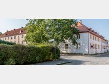 Straßenecke mit zwei Gebäuden im traditionellen Baustil, eines in gelber Putzfassade mit rotem Ziegeldach, das andere in weißer Fassade mit gelben Fensterrahmen. Im Vordergrund stehen Bäume und eine Hecke, gepflasterte Straßen und ein Straßenschild sind sichtbar.