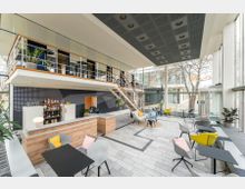 Ein modernes Café mit doppelter Deckenhöhe, großen Glasfenstern und viel Tageslicht. Im Vordergrund befinden sich Sitzbereiche mit bunten Stühlen und Tischen, im Hintergrund eine Bar mit einer Holztheke und eine Metalltreppe, die zu einer offenen Galerieebene führt.